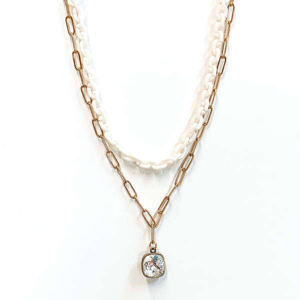 Two strand chain necklace. The shorter chain is a thick white chain. The longer chain is a simple gold paperclip chain with a hanging clear cushion cut crystal pendant with a bronze backing. This necklace is pictured on a white background.