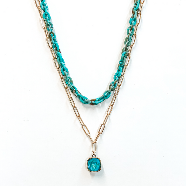 Two strand chain necklace. The shorter chain is a thick turquoise chain. The longer chain is a simple gold paperclip chain with a hanging clear cushion cut crystal pendant with a bronze backing. This necklace is pictured on a white background.