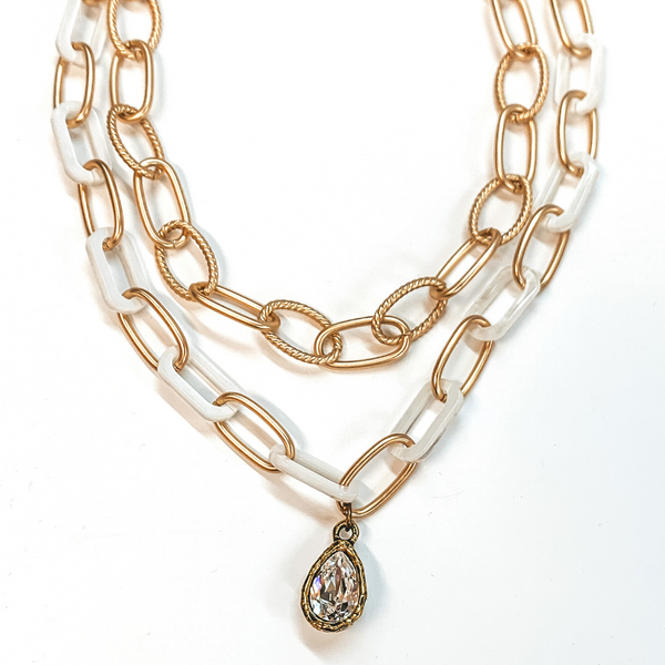 Two strand chain necklace. The shorter chain is a thick gold chain with every other chain being a wisted chain. The has gold and thick white chain links with a hanging clear teardrop crystal pendant with a bronze backing. This necklace is pictured on a white background. 