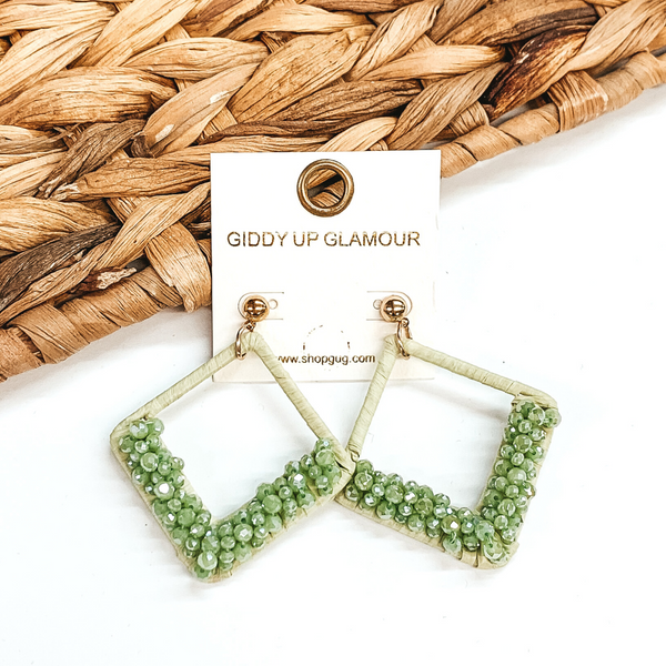 Open diamond shaped dangle earrings wrapped in raffia that includes a layer of beads on the bottom half of the earrings. These earrings are olive in color. These earrings are pictured on on a white background with basket weave decor.