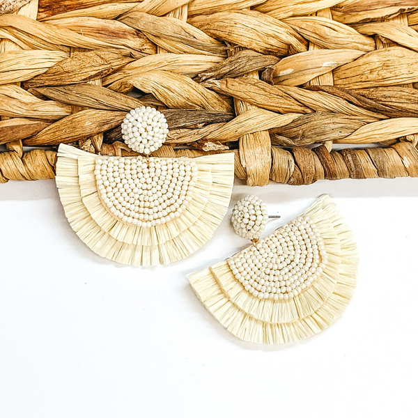 Circle beaded stud earings with a half circle beaded pendant with raffia fringe around the edge. These earrings are ivory in color. These earrings are pictured on a white background with one of the earrings leaning on tan basket weave material.