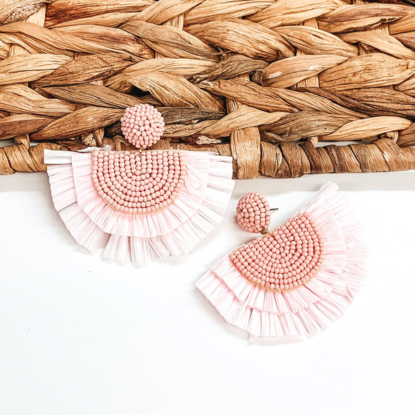 Circle beaded stud earings with a half circle beaded pendant with raffia fringe around the edge. These earrings are baby pink in color. These earrings are pictured on a white background with one of the earrings leaning on tan basket weave material.