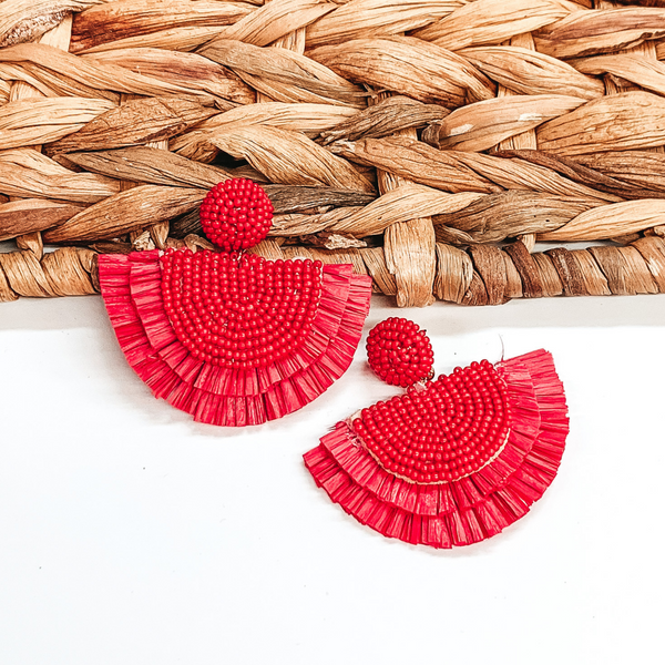 Circle beaded stud earings with a half circle beaded pendant with raffia fringe around the edge. These earrings are red in color. These earrings are pictured on a white background with one of the earrings leaning on tan basket weave material.