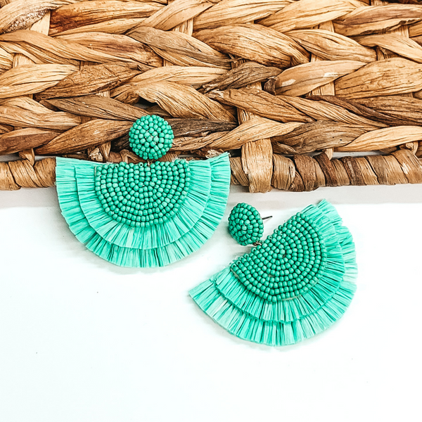 Circle beaded stud earings with a half circle beaded pendant with raffia fringe around the edge. These earrings are turquoise in color. These earrings are pictured on a white background with one of the earrings leaning on tan basket weave material.