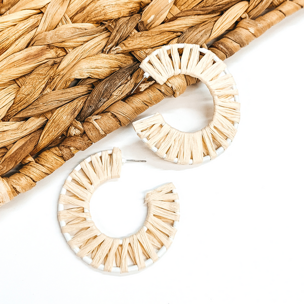 White wire circle hoop earrings that are wrapped in ivory raffia. These earrings are pictured on a white background with one of the hoops leaning on tan basket weave material.