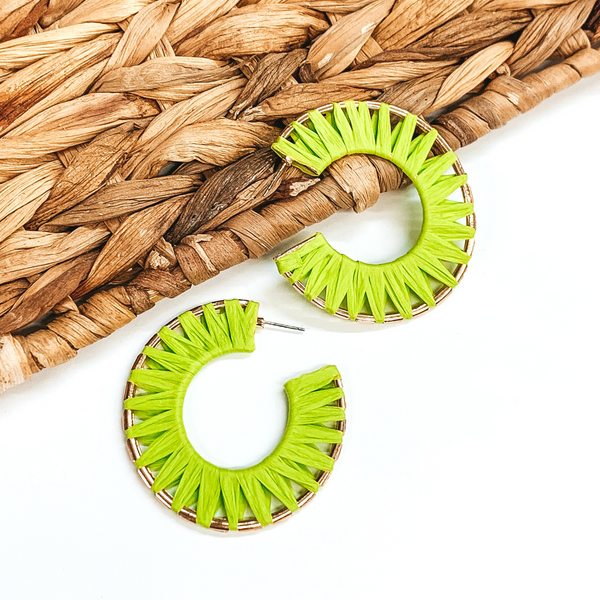 Gold wire circle hoop earrings that are wrapped in lime green raffia. These earrings are pictured on a white background with one of the hoops leaning on tan basket weave material.