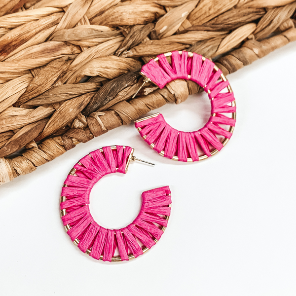 Gold wire circle hoop earrings that are wrapped in fuchsia raffia. These earrings are pictured on a white background with one of the hoops leaning on tan basket weave material.