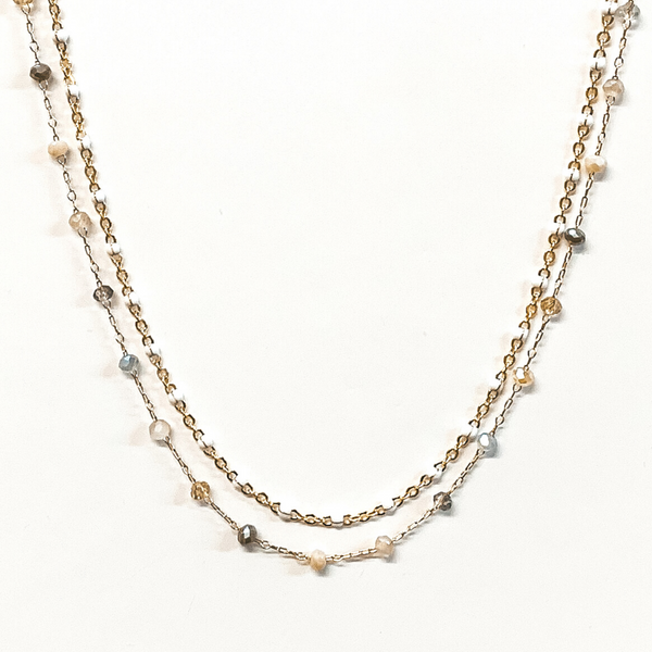 The first strand is a gold chain with small, white bead spacers. The second gold chain has small, crystal bead spacers in a mix of nude colors. This necklace is pictured on a white background.