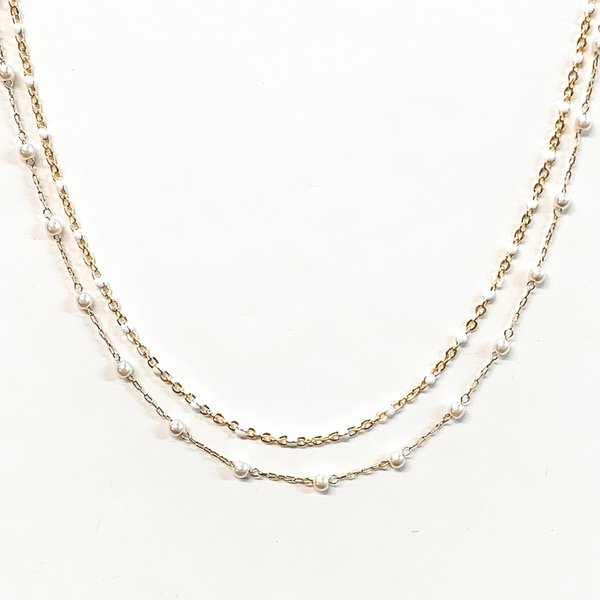 The first strand is a gold chain with small, white bead spacers. The second gold chain has small, white pearl bead spacers. This necklace is pictured on a white background.