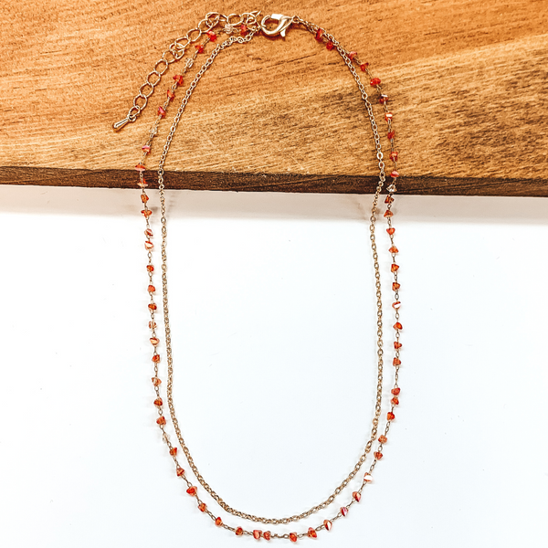 The first strand is a thin, plain gold chain. The second gold chain has small, irregular shaped crystal bead spacers in a rust. This necklace is pictured laying partially on a wood block on a white background.