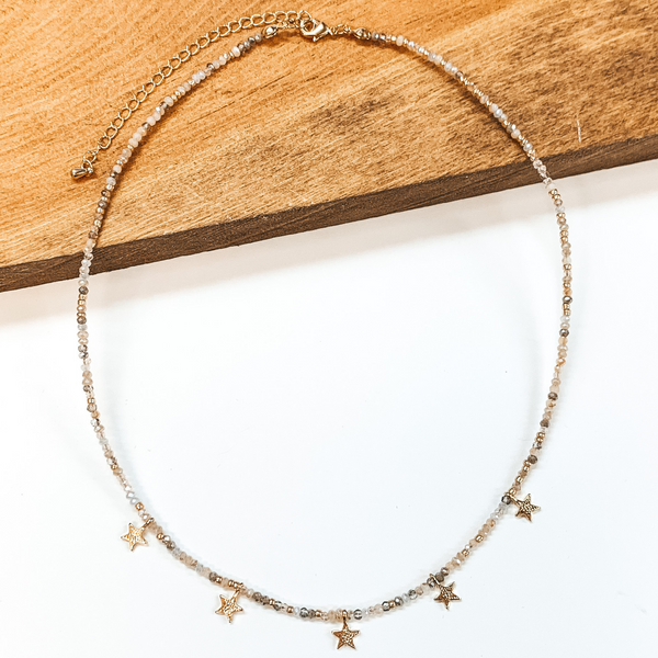 This is a beaded necklace with gold bead spacerss and five gold star charms. This neckalce includes a nude mix of beads. This necklace is pictured laying partially on a wood block on a white background.