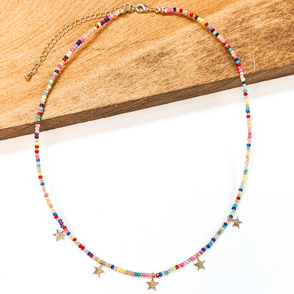 This is a beaded necklace with gold bead spacerss and five gold star charms. This neckalce includes a neon mix of beads. This necklace is pictured laying partially on a wood block on a white background.