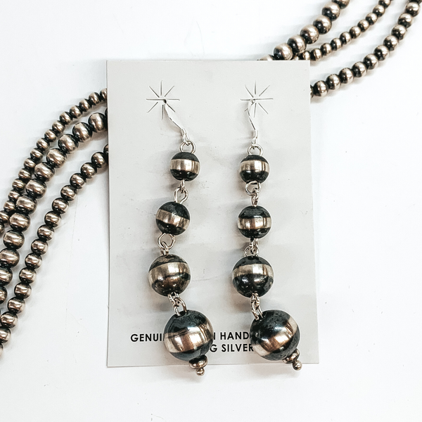 Silver, graduated beaded earrings. These earrings are pictured on a white background on top of silver beads. 