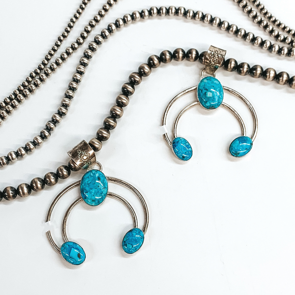 Two silver naja pendants with silver bails and a center turquoise stone and turquoise stones at the ends. This pendant is pictured on a white background with silver beads above the pendant.