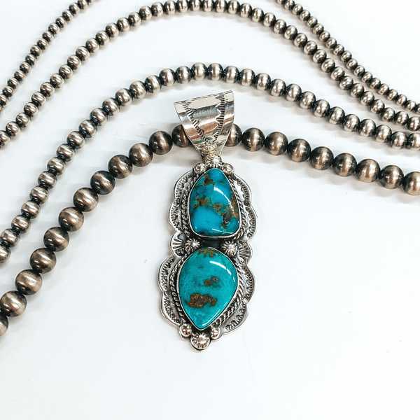 Silver, detailed bail with a connecting asymmetrical shaped pendant. This pendant includes engraved detailing and two turquoise stones. This pendant is pictured on a white background with silver beads above the pendant. 