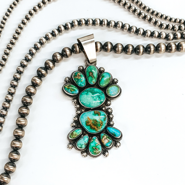Silver bail with turquoise stone cluster pendant attached. This pendant includes 12 turquoise stones and silver detailed beads outlining the stones. This pendant is pictured on a white background with silver beads above the pendant. 
