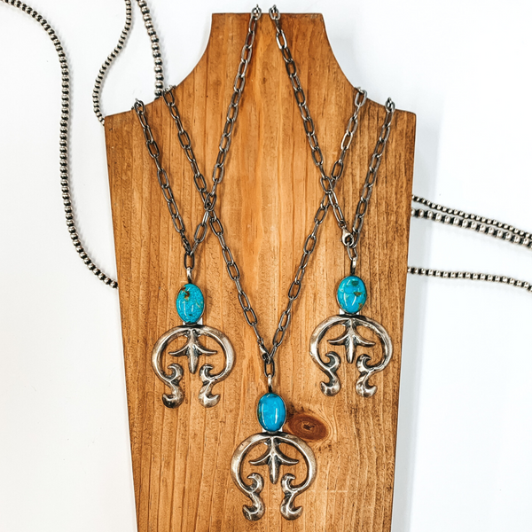 Silver paperclip chain with a connecting oval turquoise stone and silver naja pendant. There are three necklaces pictured on a brown necklace holder on a white background. 