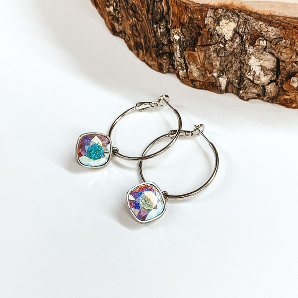 Two silver hoop earrings with a square AB crystal. These earrings are pictured on a white background with a piece of wood at the top.