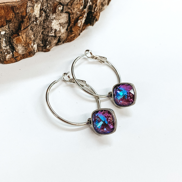 Two silver hoop earrings with a square burgundy delight crystal. These earrings are pictured on a white background with a piece of wood at the top.