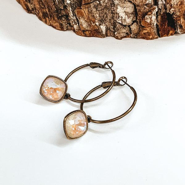 Two bronze hoop earrings with a square ivory cream delight crystal. These earrings are pictured on a white background with a piece of wood at the top.