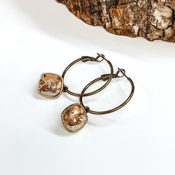 Two bronze hoop earrings with a square golden shadow crystal. These earrings are pictured on a white background with a piece of wood at the top.