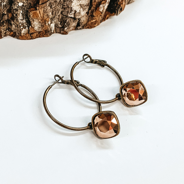 Two bronze hoop earrings with a square rose gold crystal. These earrings are pictured on a white background with a piece of wood at the top.