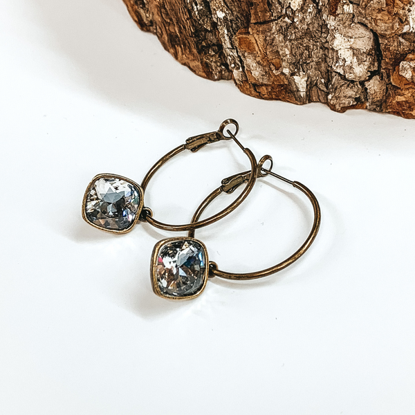 Two bronze hoop earrings with a square crystal ignite crystal. These earrings are pictured on a white background with a piece of wood at the top.