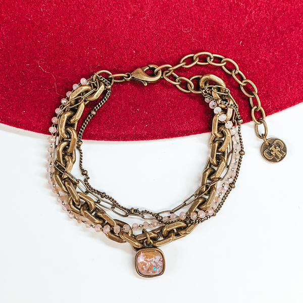 Bronze, adjustable bracelet that includes a thick chain strnad, crystal beaded strand, a thin paperlcip chain strand, and a small chain strand. There is also a cappuccino delight cushion cut crystal drop. this bracelet is pictured on a white and red background. 