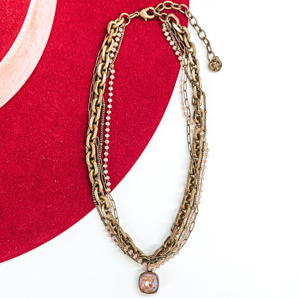 Bronze, adjustable necklace that includes a thick chain strand, crystal beaded strand, a thin paperlcip chain strand, and a small chain strand. There is also a cappuccino delight cushion cut crystal drop. This necklace is pictured on a white and red background.