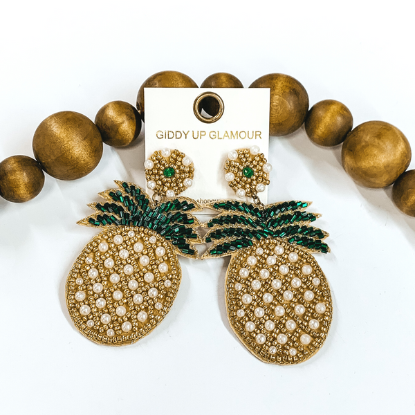 Circle beaded stud earrings with a beaded pineapple. These earrings include pearl, gold, and green beads. These earrings are pictured on a white background with brown beads behind them. 