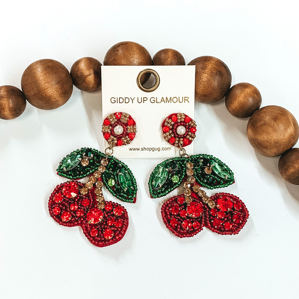 Circle beaded stud earrings with beaded cherries. These earrings include red and green beads with red, green, and bronze crystals. These earrings are pictured on a white background with brown beads behind them. 