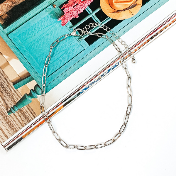 Silver paperclip chain necklace that is pictured half on a magazine and half on a white background. 