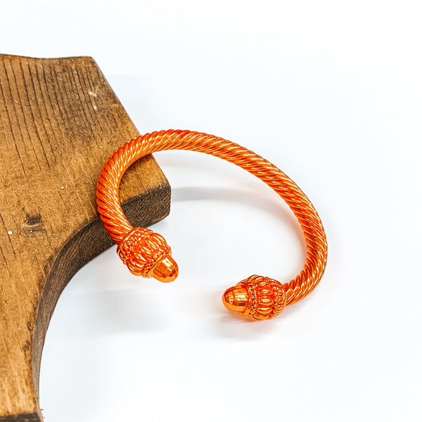 Cable bracelet with big cabochon ends in metallic orange. This bracelet is pictured on a white background and leaning on a piece of brown wood. 