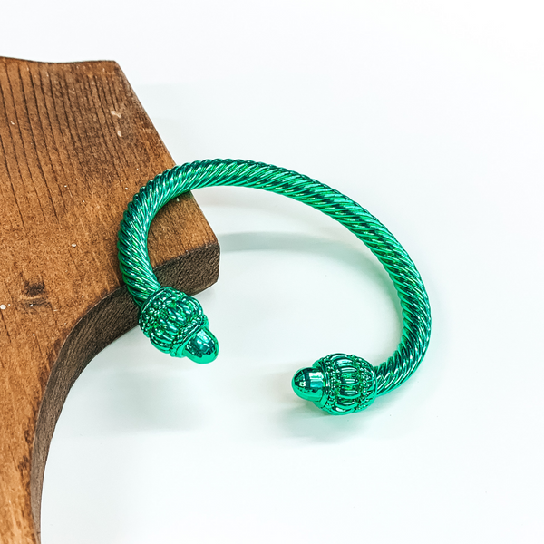 Cable bracelet with big cabochon ends in metallic green. This bracelet is pictured on a white background and leaning on a piece of brown wood.