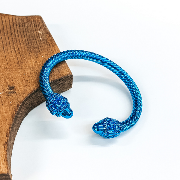 Cable bracelet with big cabochon ends in metallic blue. This bracelet is pictured on a white background and leaning on a piece of brown wood.  