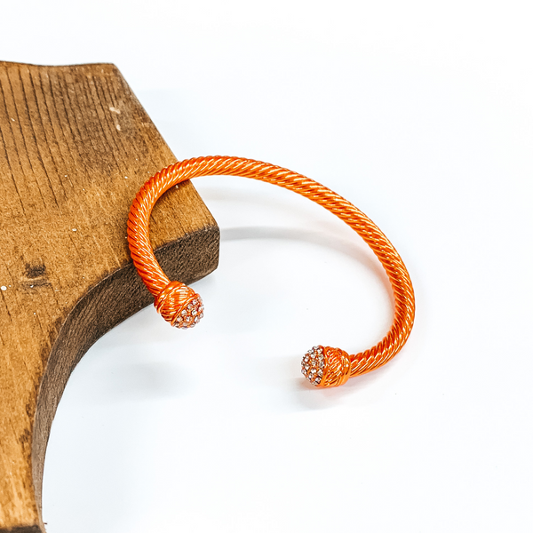 Cable bracelet with clear crystal cabochon ends in metallic orange. This bracelet is pictured on a white background and leaning on a piece of brown wood. 