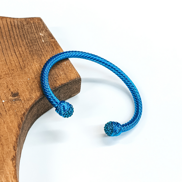 Cable bracelet with clear crystal cabochon ends in metallic blue. This bracelet is pictured on a white background and leaning on a piece of brown wood.  