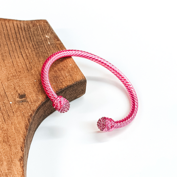 Cable bracelet with clear crystal cabochon ends in metallic pink. This bracelet is pictured on a white background and leaning on a piece of brown wood. 