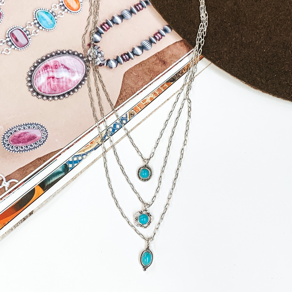 Triple layered, silver, chain necklaces. each necklace has a small turquoise stone pendant with a silver outlining. This necklace is pictured partially on an open magazine on a white background. 