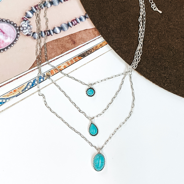 Triple layered, silver, chain necklaces. Each necklace has a small turquoise stone pendant with a silver outlining. This necklace is pictured partially on an open magazine on a white background. 