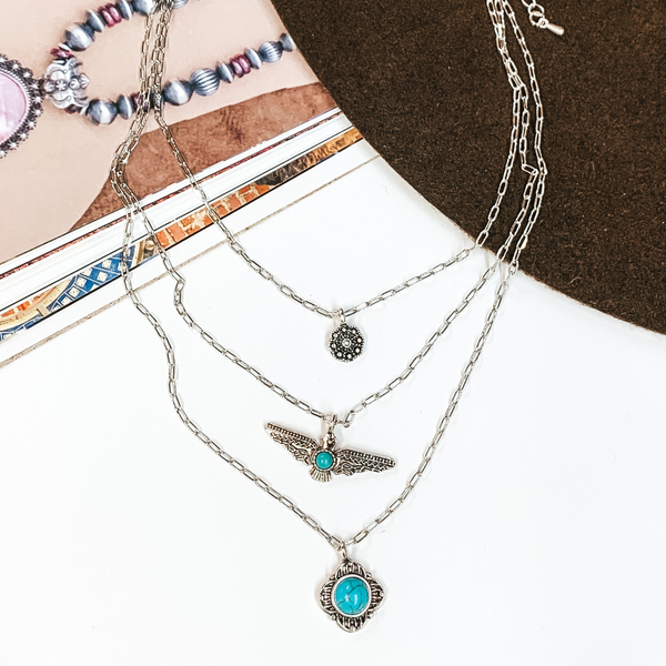 Triple layered, silver, chain necklaces. Each necklace has a pendant which inlcudes a silver flower concho, a silver thunderbird with center turquoise stone, and a circle turquoise stone with silver outline. This necklace is pictured partially on an open magazine on a white background. 