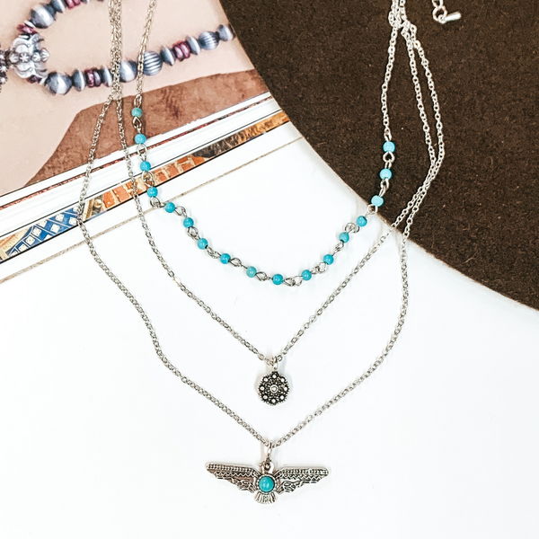 Triple layered, silver, chain necklaces. Two strands have a pendant which inlcudes a silver flower concho and a silver thunderbird with center turquoise stone, and the shortest chain has a turquoise beaded section. This necklace is pictured partially on an open magazine on a white background.