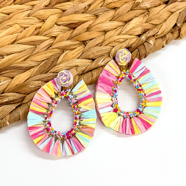 Circle beaded earrings with a hanging open teardrop pendant. The pendant is a beaded teardrop with raffia wrapped around the edges. The earrings are pastel multicolored. These earrings are pictured on a white backround while partially laying on a tan woven material.