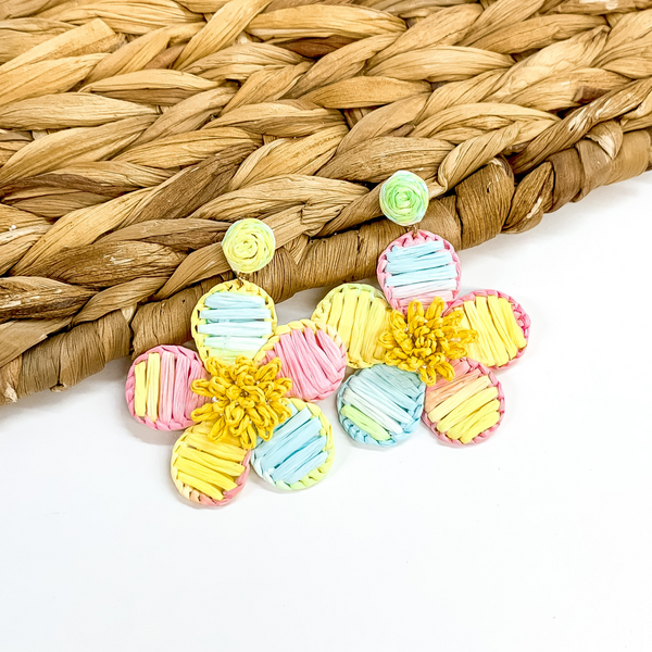 Pastel multicolored affia wrapped circle studs with hanging flower pendants wrapped in pastel multicolored raffia. These earrings are pictured partially laying on a basket weave material on a white background.
