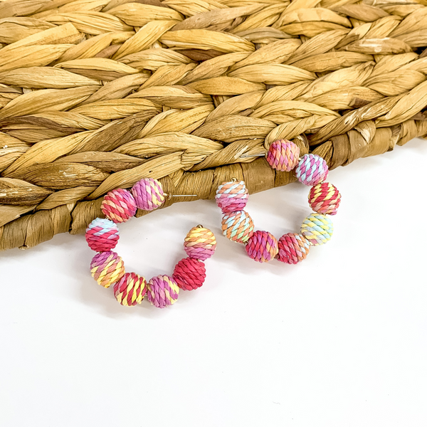 Hoop earrings with raffia balls around. The balls are wrapped in pastel multicolored raffia. Taken on a white background and partially laying on a woven basket material.
