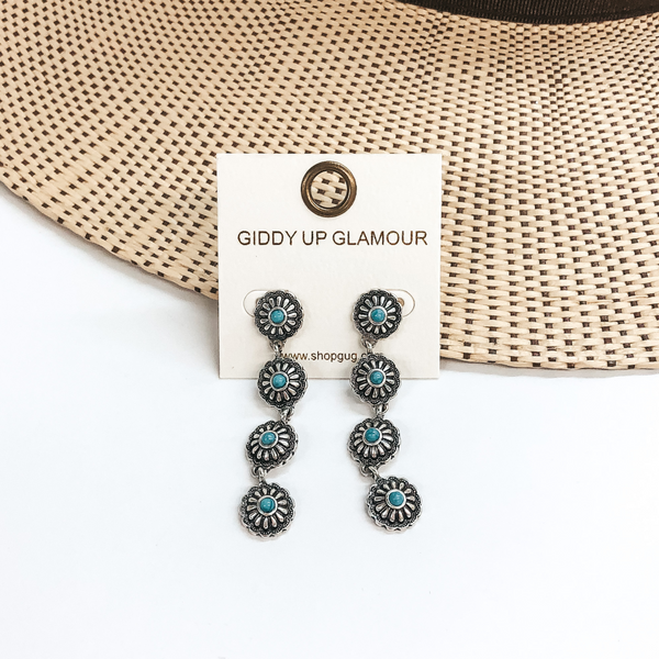 Four small, silver conchos in a row to form a dangle earrings. Each concho has a small turquoise stone in the center. These earrings are pictured on a white and tan background. 