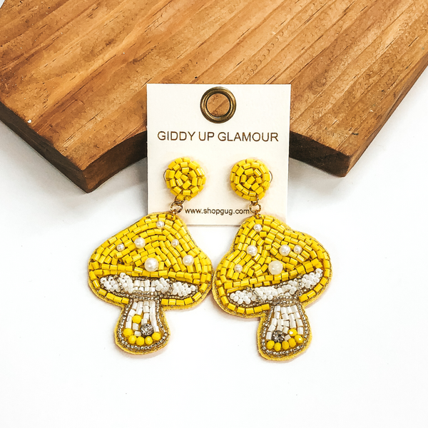 Yellow beaded circle studs with a hanging yellow beaded mushroom pendant. The mushroom also includes some white beaded detailing and some white pearls. these earrings are pictured in front of a wood block on a white background. 