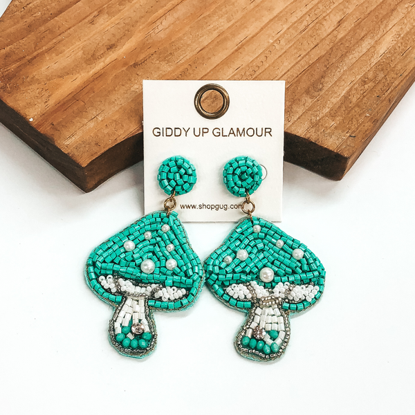 Turquoise beaded circle studs with a hanging turquoise beaded mushroom pendant. The mushroom also includes some white beaded detailing and some white pearls. these earrings are pictured in front of a wood block on a white background. 