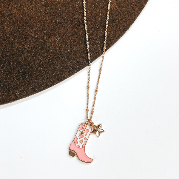 Gold chain necklace with a gold star charm and a white and pink cow print boot charm. This necklace is pictured on a white and brown background. 