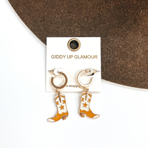Small gold hoop earrings with a ivory and tan boot pendant hanging from the bottom. These earrings are pictured on a white and brown background.